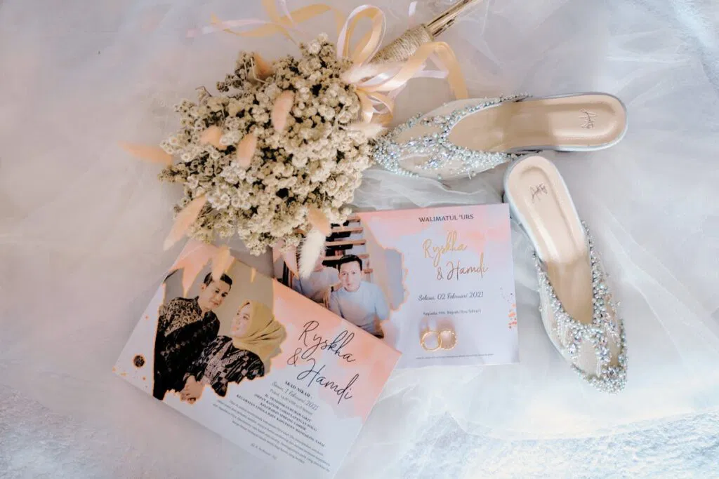 Wedding invitations, a pair of bridal shoes, rings, and a bouquet on a fabric surface with Christmas cards.