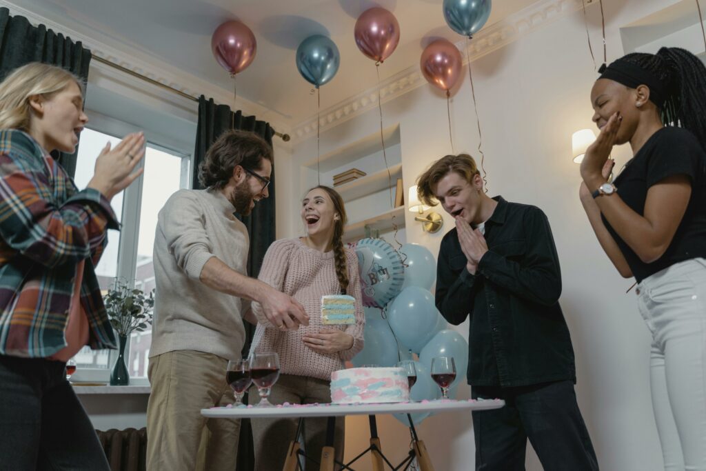 A group of four people stands around a table with a cake. Two hold cake slices, while the others react with joyful expressions. Balloons and drinks set the festive scene, which was captured in party photos with printing on site for instant memories.