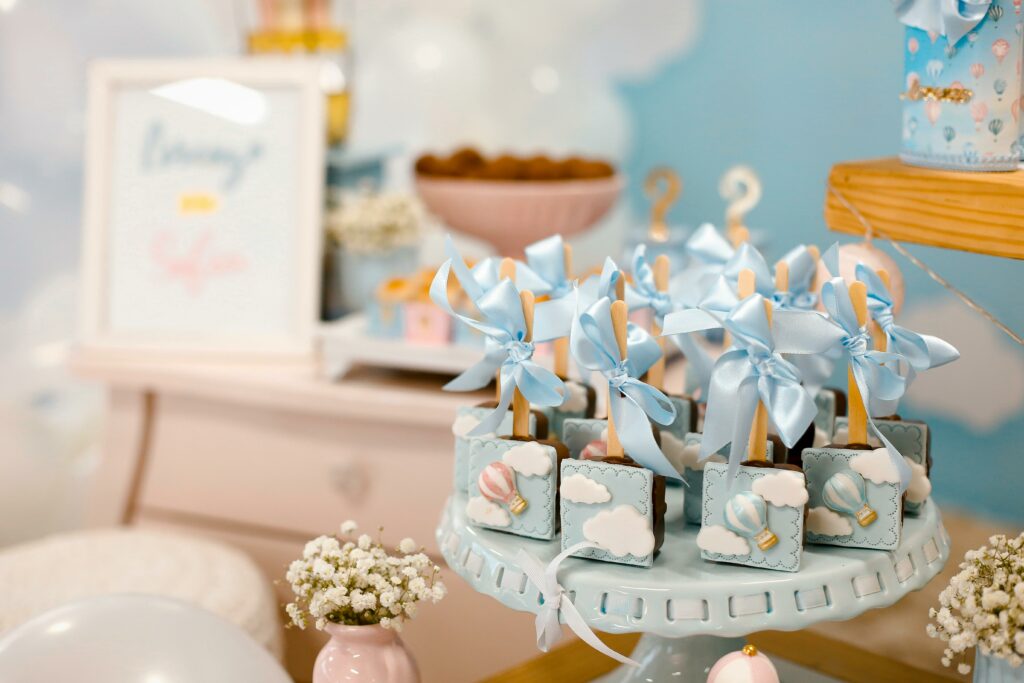 7 - Personal Touch Printing A display of baby shower-themed treats featuring blue ribbon-tied chocolates with hot air balloon designs. The background includes balloons, flowers, and a soft pastel setup. Personal touch printing adds party photos with printing on site to capture and cherish the precious moments.