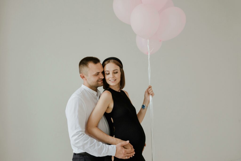 pexels-rdne-9214930 - Personal Touch Printing A man embraces a pregnant woman who is holding a bunch of pink balloons against a plain background, both with calm expressions. The tender moment captured could beautifully adorn Memorial Programs-Posters-DVDs for an unforgettable keepsake.